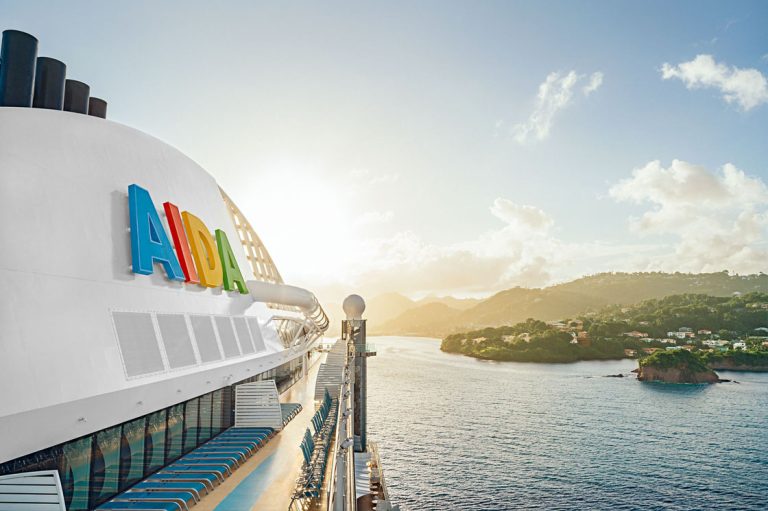 Blick vom Deck eines AIDA Kreuzfahrtschiffs bei Sonnenaufgang, mit farbigem AIDA-Logo im Vordergrund und tropischer Küstenlandschaft im Hintergrund.