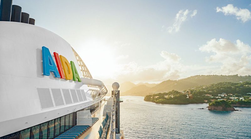 Blick vom Deck eines AIDA Kreuzfahrtschiffs bei Sonnenaufgang, mit farbigem AIDA-Logo im Vordergrund und tropischer Küstenlandschaft im Hintergrund.