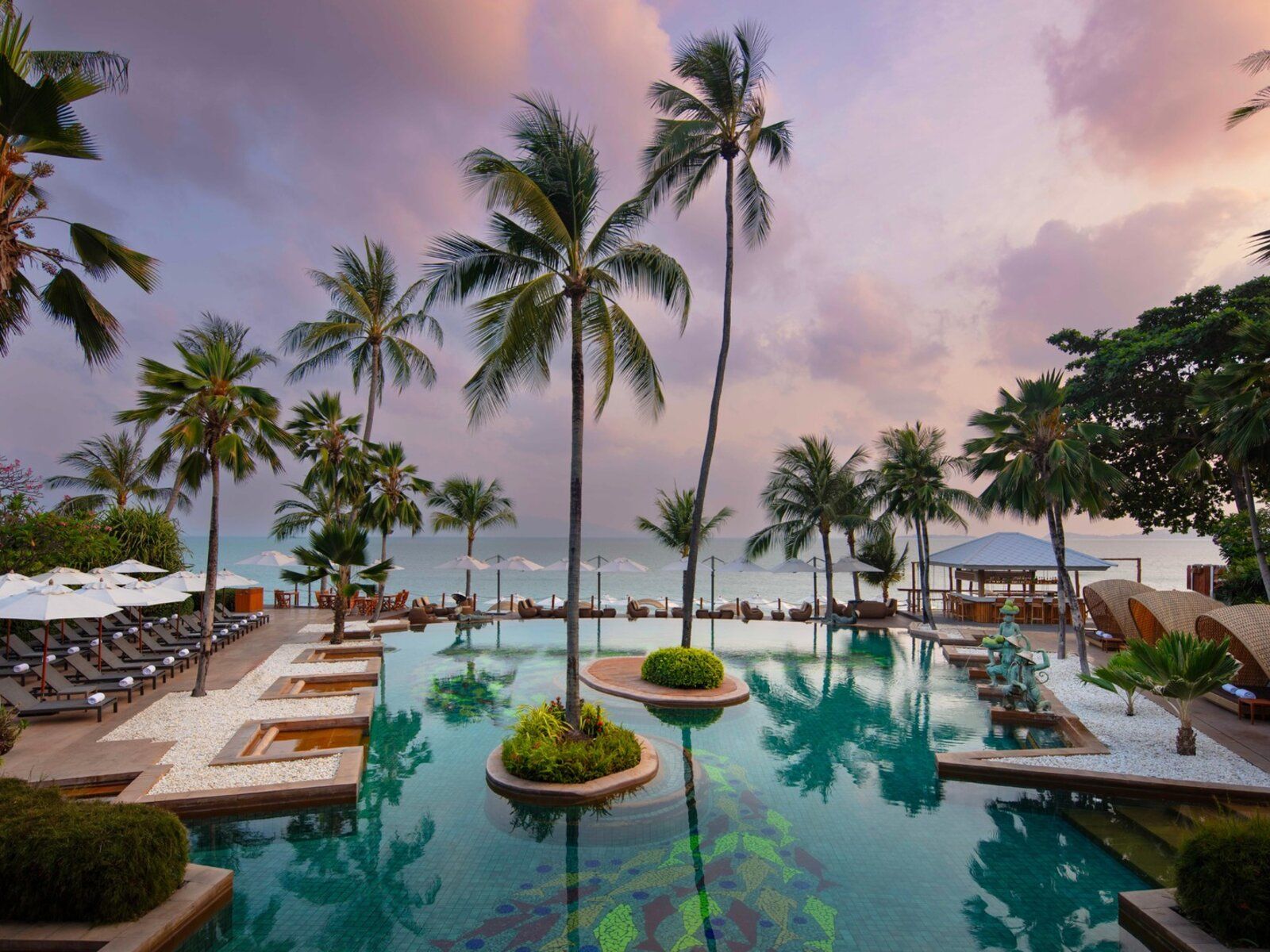 Traumhafter Poolbereich im Anantara Bophut Koh Samui Resort bei Sonnenuntergang – von Palmen gesäumt, mit luxuriösen Liegen und direktem Blick auf den weißen Sandstrand und das ruhige, türkisfarbene Meer. Eine stilvolle Strandbar und kunstvolle Wasserelemente sorgen für eine exotische Atmosphäre.