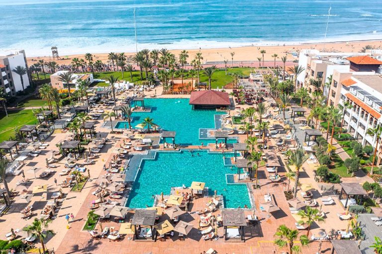 Ein luxuriöses Resort mit einem großen, geometrisch gestalteten Poolbereich direkt am Sandstrand. Zahlreiche Sonnenliegen, Sonnenschirme und balinesische Betten umgeben die Pools, während tropische Palmen und grüne Gärten eine entspannte Atmosphäre schaffen. Im Hintergrund erstreckt sich das blaue Meer mit sanften Wellen, und ein Holzsteg führt zum Strand, wo Menschen spazieren gehen. Die Hotelgebäude im modernen Stil mit weißen Fassaden und Terrakotta-Dächern rahmen das Bild harmonisch ein.
