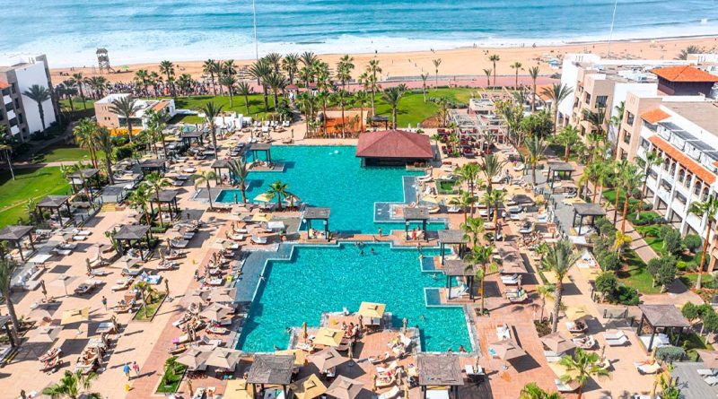 Ein luxuriöses Resort mit einem großen, geometrisch gestalteten Poolbereich direkt am Sandstrand. Zahlreiche Sonnenliegen, Sonnenschirme und balinesische Betten umgeben die Pools, während tropische Palmen und grüne Gärten eine entspannte Atmosphäre schaffen. Im Hintergrund erstreckt sich das blaue Meer mit sanften Wellen, und ein Holzsteg führt zum Strand, wo Menschen spazieren gehen. Die Hotelgebäude im modernen Stil mit weißen Fassaden und Terrakotta-Dächern rahmen das Bild harmonisch ein.