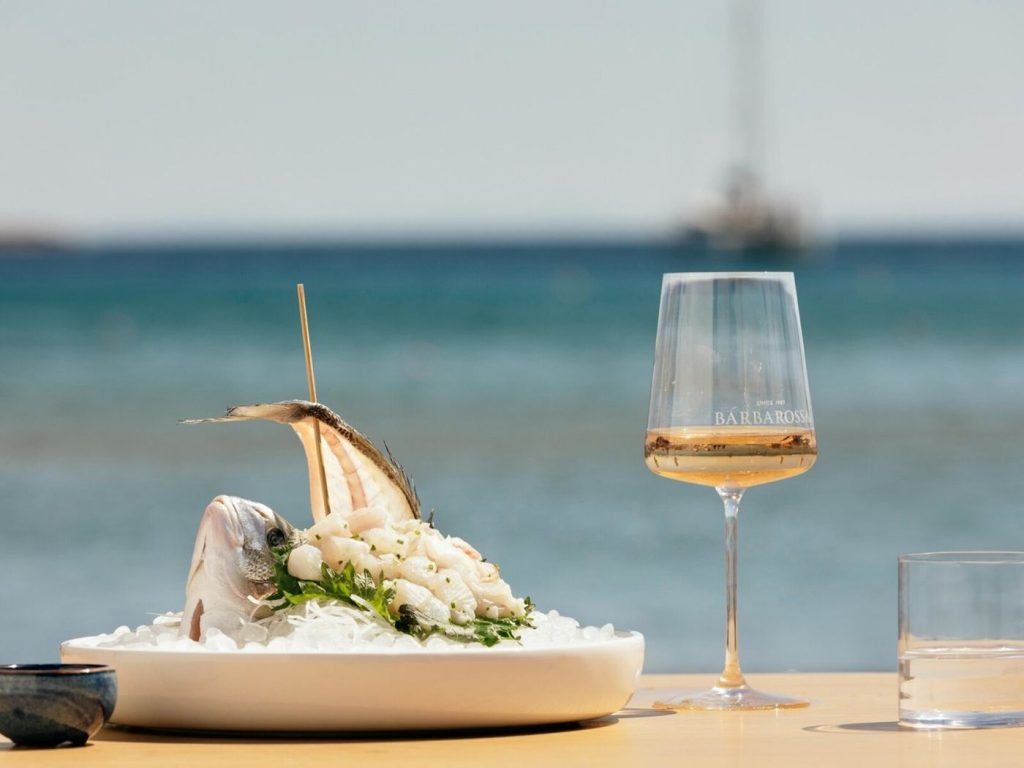 Ein kunstvoll angerichtetes Sashimi-Gericht mit Fisch und Meeresfrüchten auf Eis, serviert neben einem Glas Roséwein mit Meeresblick im Hintergrund.