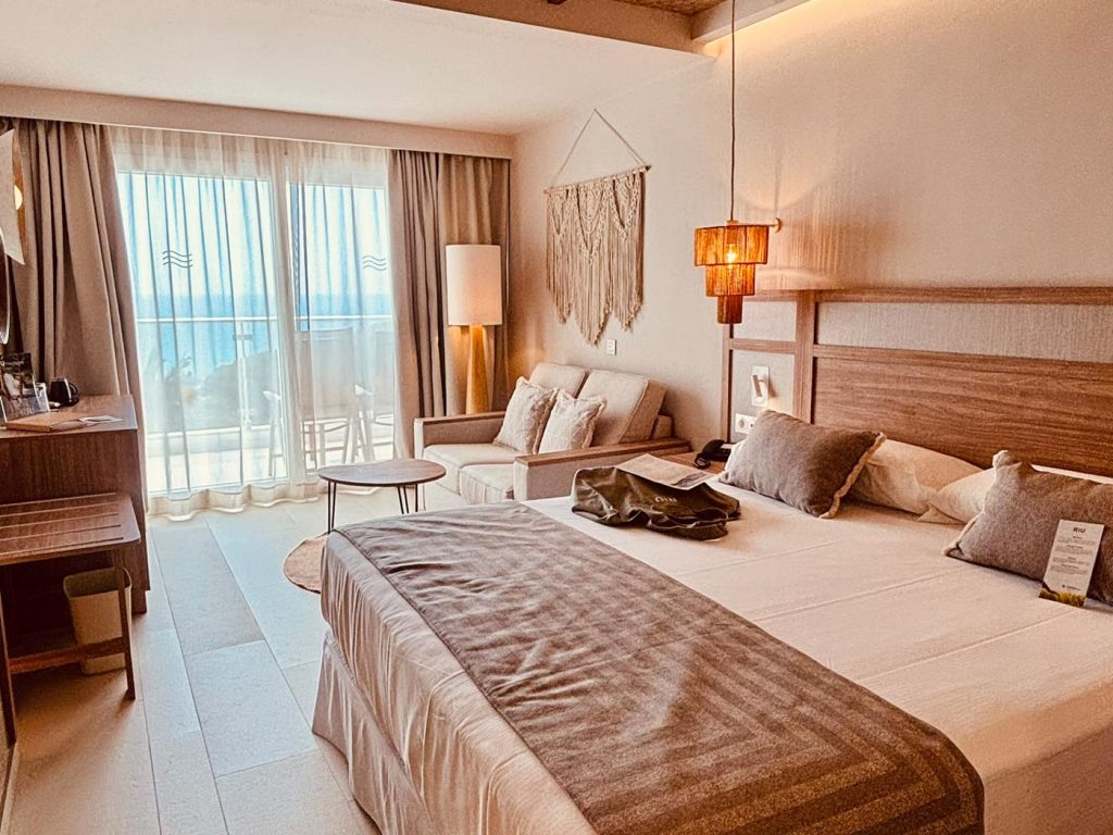 Modern eingerichtetes Hotelzimmer mit Doppelbett, Sofa, Naturtönen, Holzelementen und Balkon mit Meerblick im Riu Palace La Mola auf Formentera.