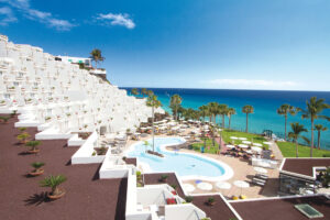 Blick auf das frisch renovierte Hotel Riu Palace Calypso auf Fuerteventura mit weißer Architektur, Poollandschaft, Palmen und Meer im Hintergrund.