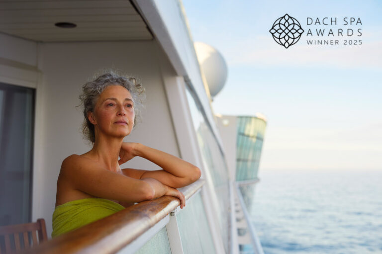 Eine Frau in einem grünen Handtuch lehnt entspannt am Balkon eines Kreuzfahrtschiffs, rechts oben das Logo „DACH Spa Awards Winner 2025“.