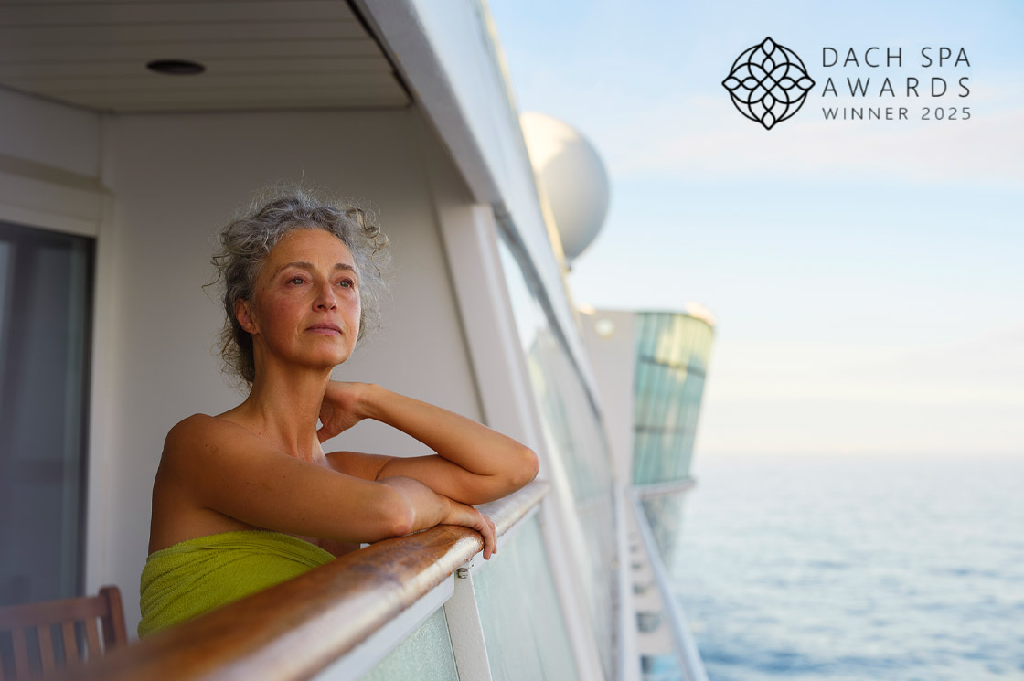 Eine Frau in einem grünen Handtuch lehnt entspannt am Balkon eines Kreuzfahrtschiffs, rechts oben das Logo „DACH Spa Awards Winner 2025“.