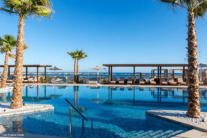 Außenpool des allsun Hotel Carolina Sun Beach mit Palmen, Sonnenliegen und Blick auf das Meer unter blauem Himmel.