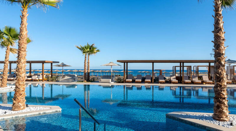 Außenpool des allsun Hotel Carolina Sun Beach mit Palmen, Sonnenliegen und Blick auf das Meer unter blauem Himmel.