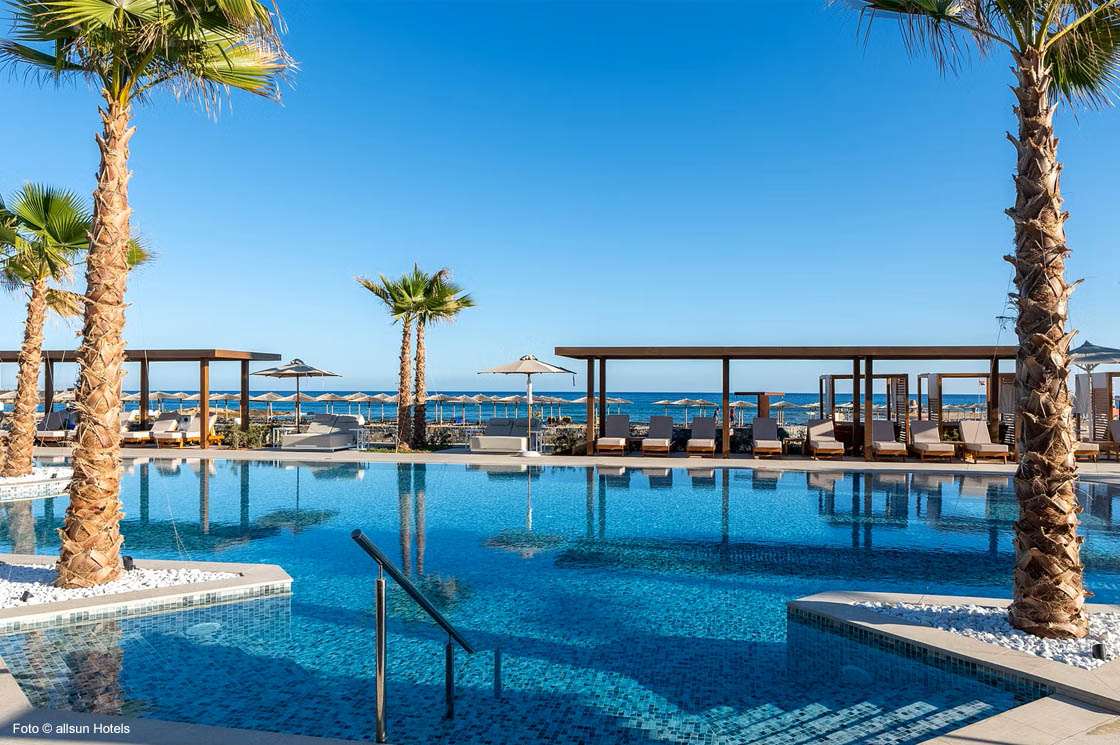 Außenpool des allsun Hotel Carolina Sun Beach mit Palmen, Sonnenliegen und Blick auf das Meer unter blauem Himmel.