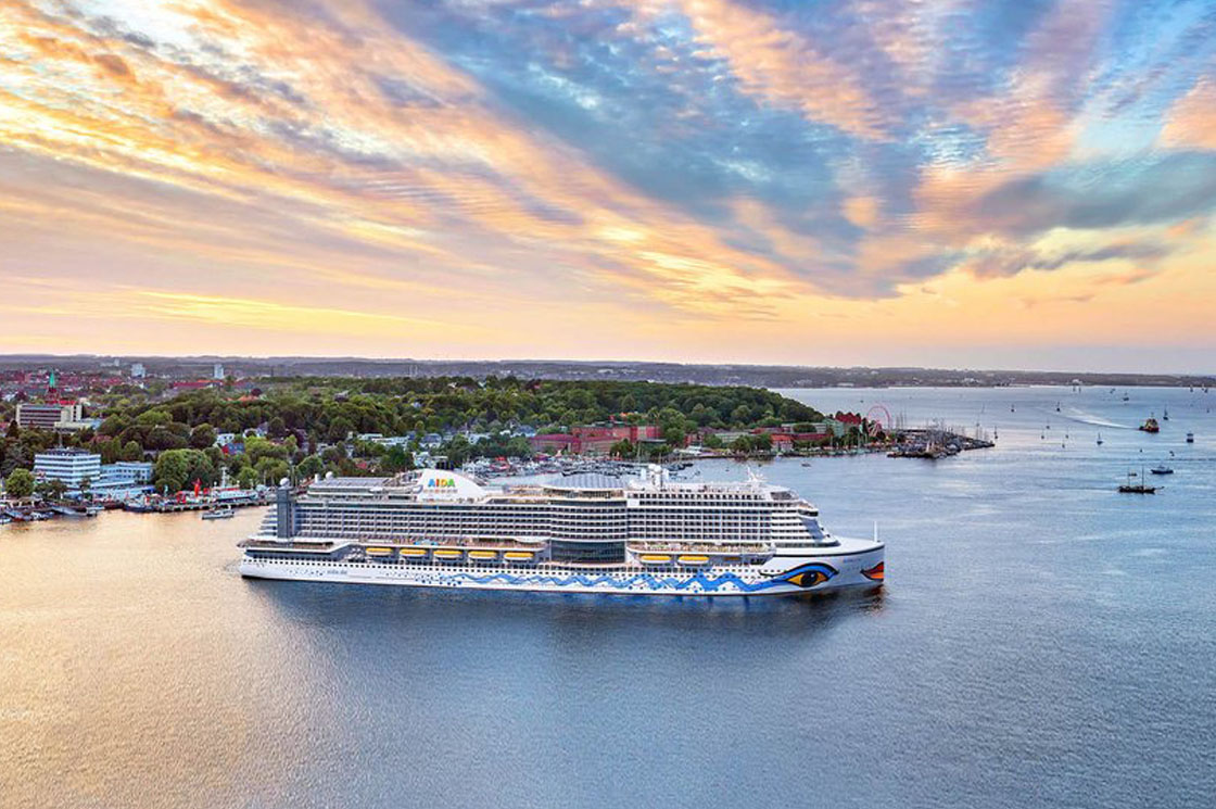 Die AIDAperla liegt vor einer Küstenstadt bei Sonnenuntergang, während sich der Himmel farbenreich über dem Wasser spiegelt.